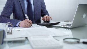 Businessman wearing blue suit is working with laptop, calculator and documents in a professional office setting.
